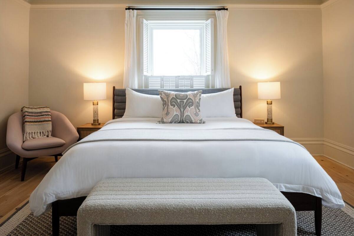A neatly arranged bedroom with a large bed, two bedside lamps, a chair, and a window with curtains.