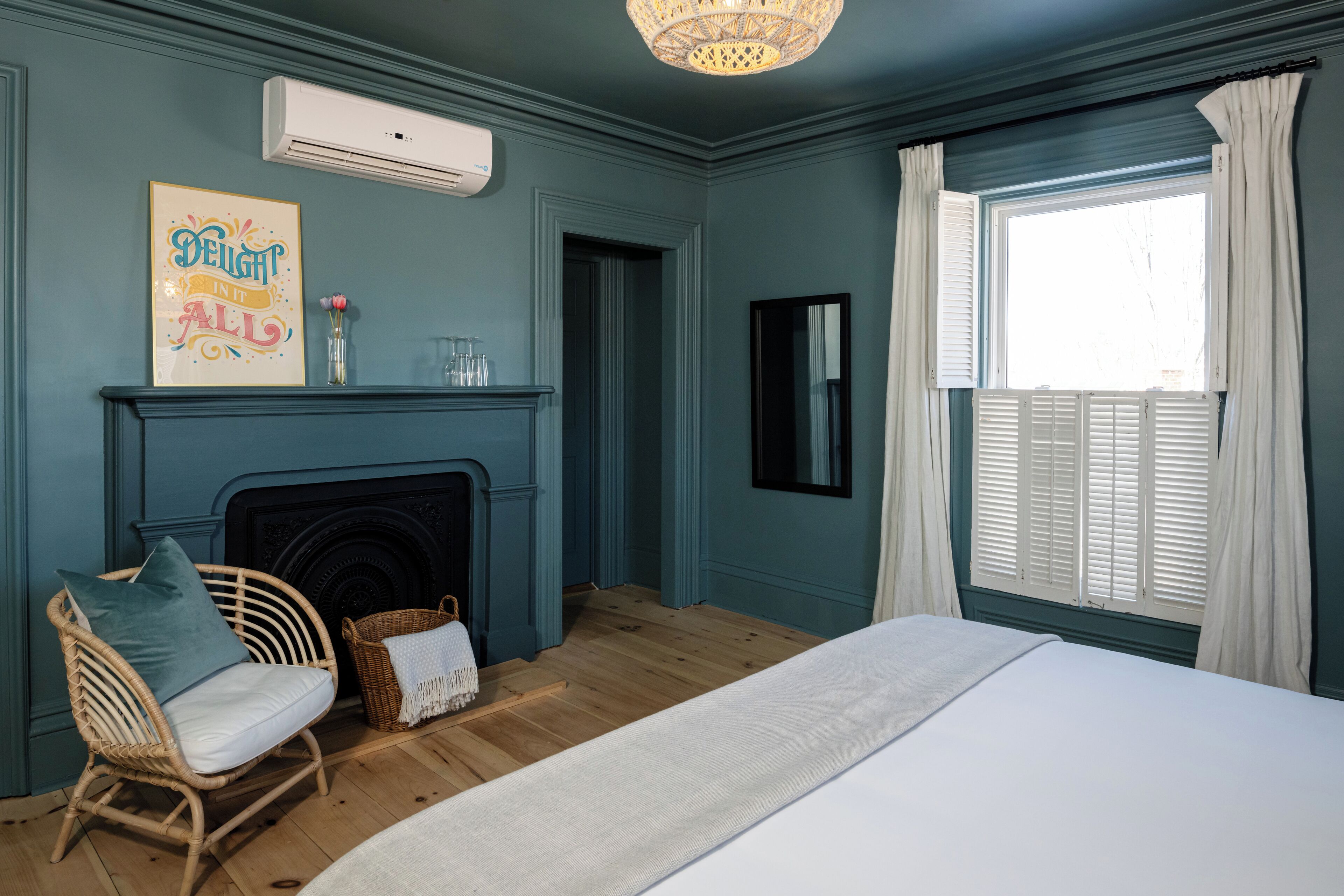 A bedroom with a bed, a wicker chair, a fireplace, a window with curtains, and a wall-mounted air conditioner.
