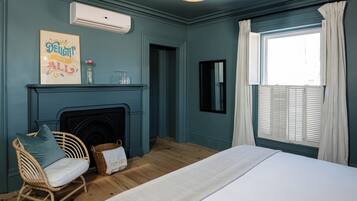 A bedroom with a bed, a wicker chair, a fireplace, a window with curtains, and a wall-mounted air conditioner.