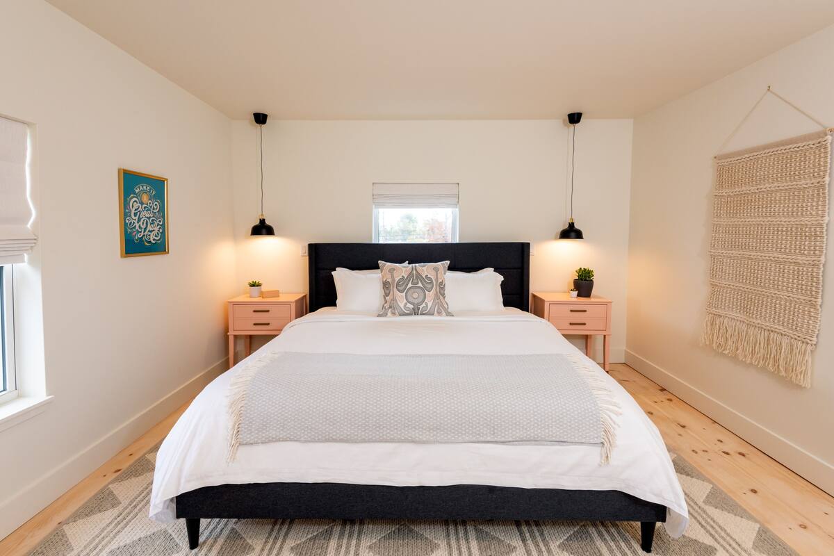 A bedroom with a large bed, bedside tables, a wall-mounted macrame piece, and a window with blinds.