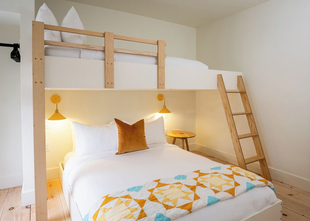 Una cama literera con escalera, una mesita de noche con lámpara y una colcha de estampado geométrico.