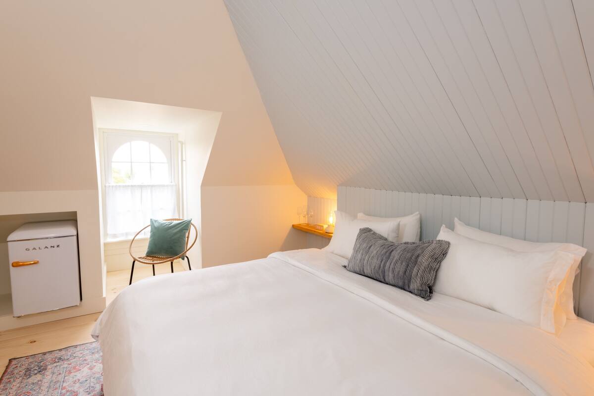 A bedroom with a large bed, a nightstand with a lamp, a chair, and a small white appliance.