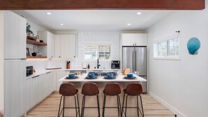 A modern kitchen with white cabinetry, a central island with bar stools, a stainless steel refrigerator, and a decorative shell on the wall. - Zanzibar Retreat 2BR | Walk to Beach! + Courtyard (San Diego)