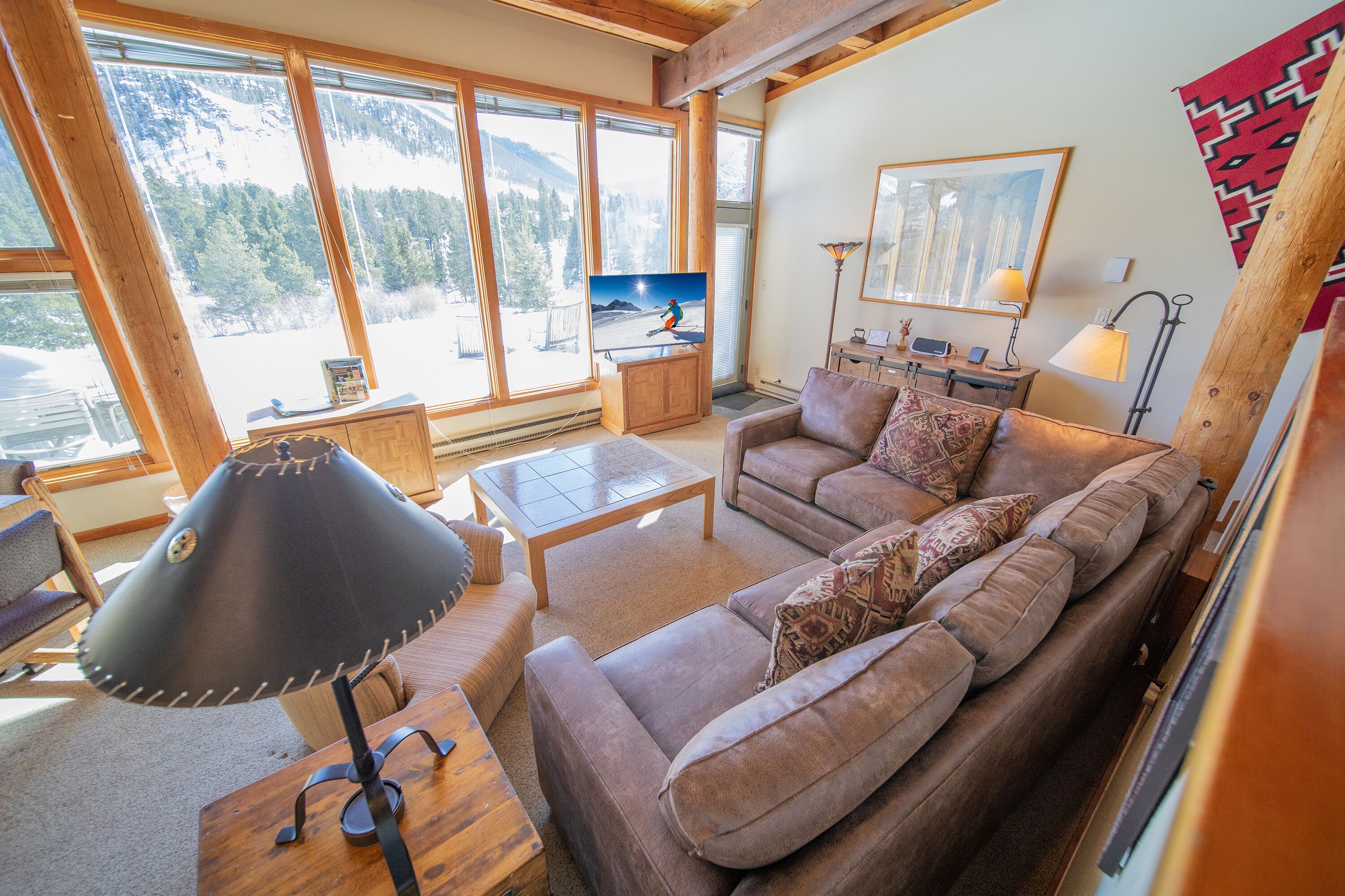 A cozy living room with a sofa, coffee table, and TV, large windows with a mountain view, and wooden beams on the ceiling.