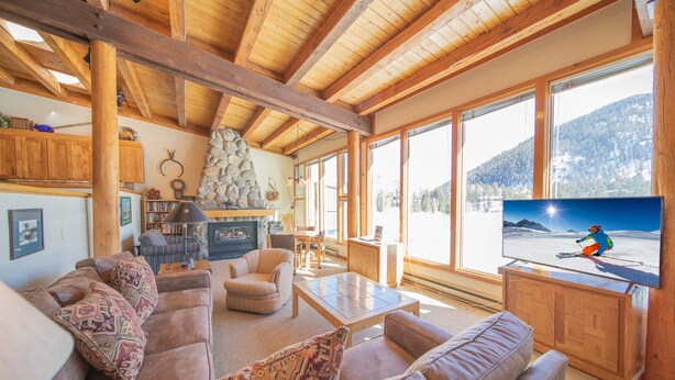 A cozy living room with a wooden ceiling, a large TV, and a view of a snowy mountain. - Homestead 2024 (Keystone)