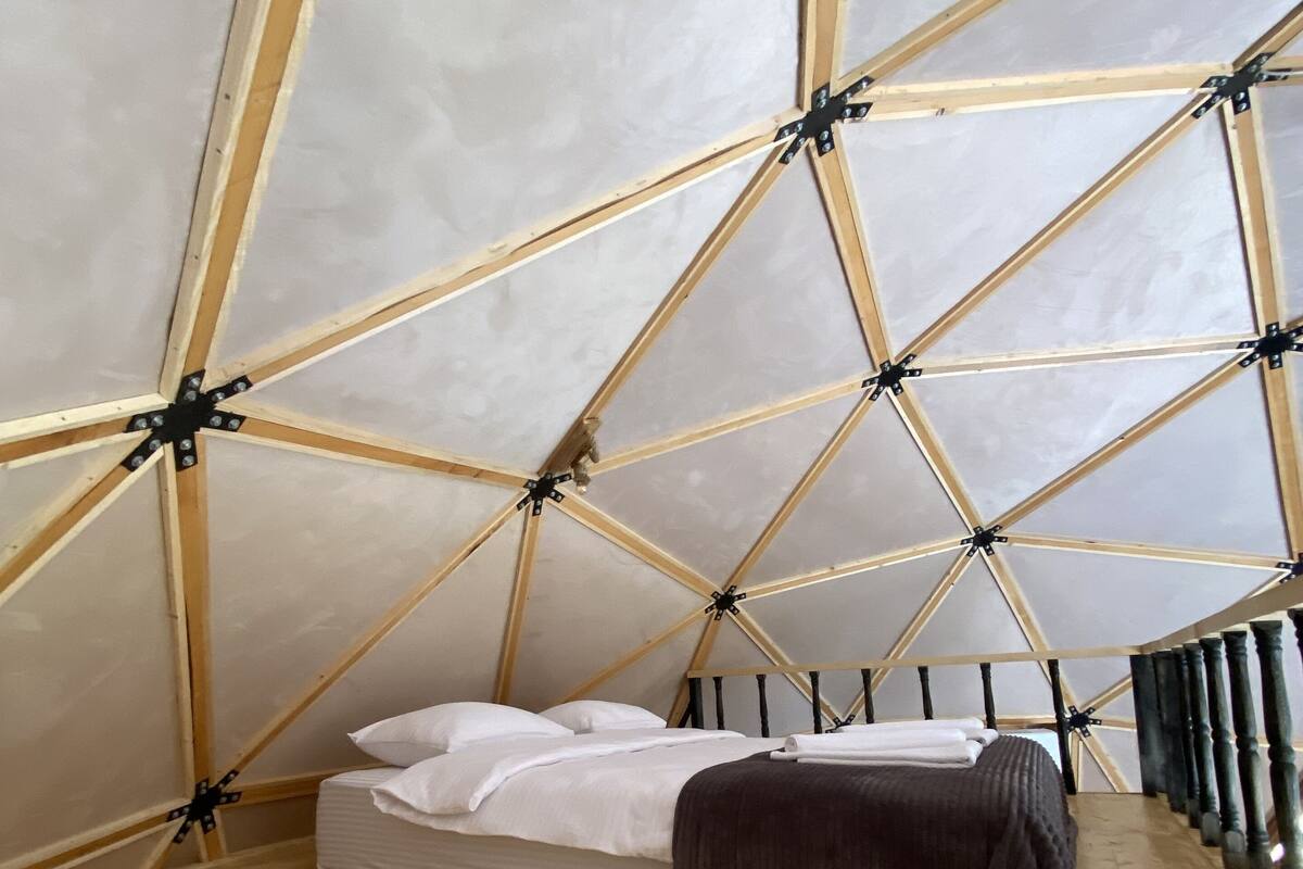 Quarto com estrutura de teto em madeira geométrica, cama com roupa de cama branca e piso de madeira.