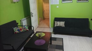 Une pièce avec un mur vert, un canapé noir, un fauteuil noir, une petite table sur laquelle est posée une nappe violette, et une porte qui donne sur une autre pièce.