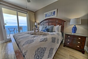 A bedroom with a large bed, a nightstand, a lamp, and a view of the ocean through a sliding door. - Ocean Breeze (Panama City Beach)