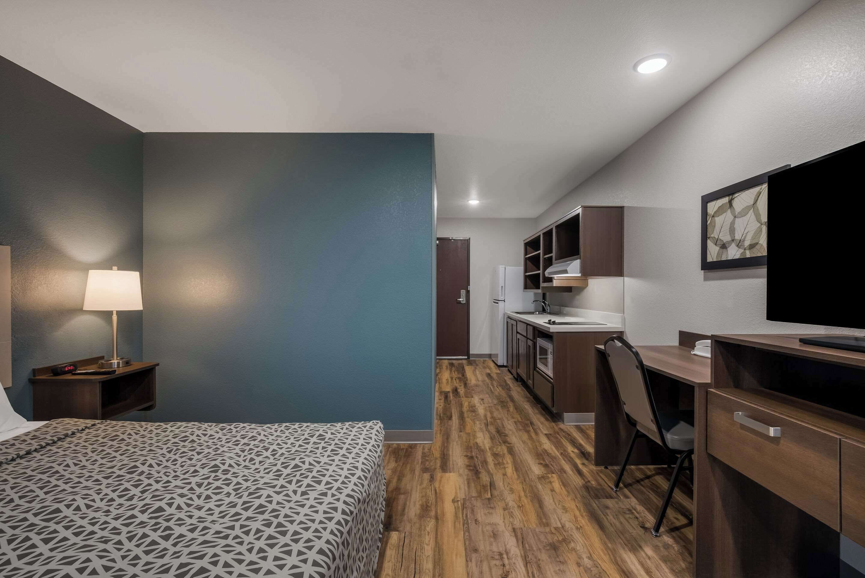 Una habitación de hotel compacta con cama, escritorio, silla, kitchenette y televisor.