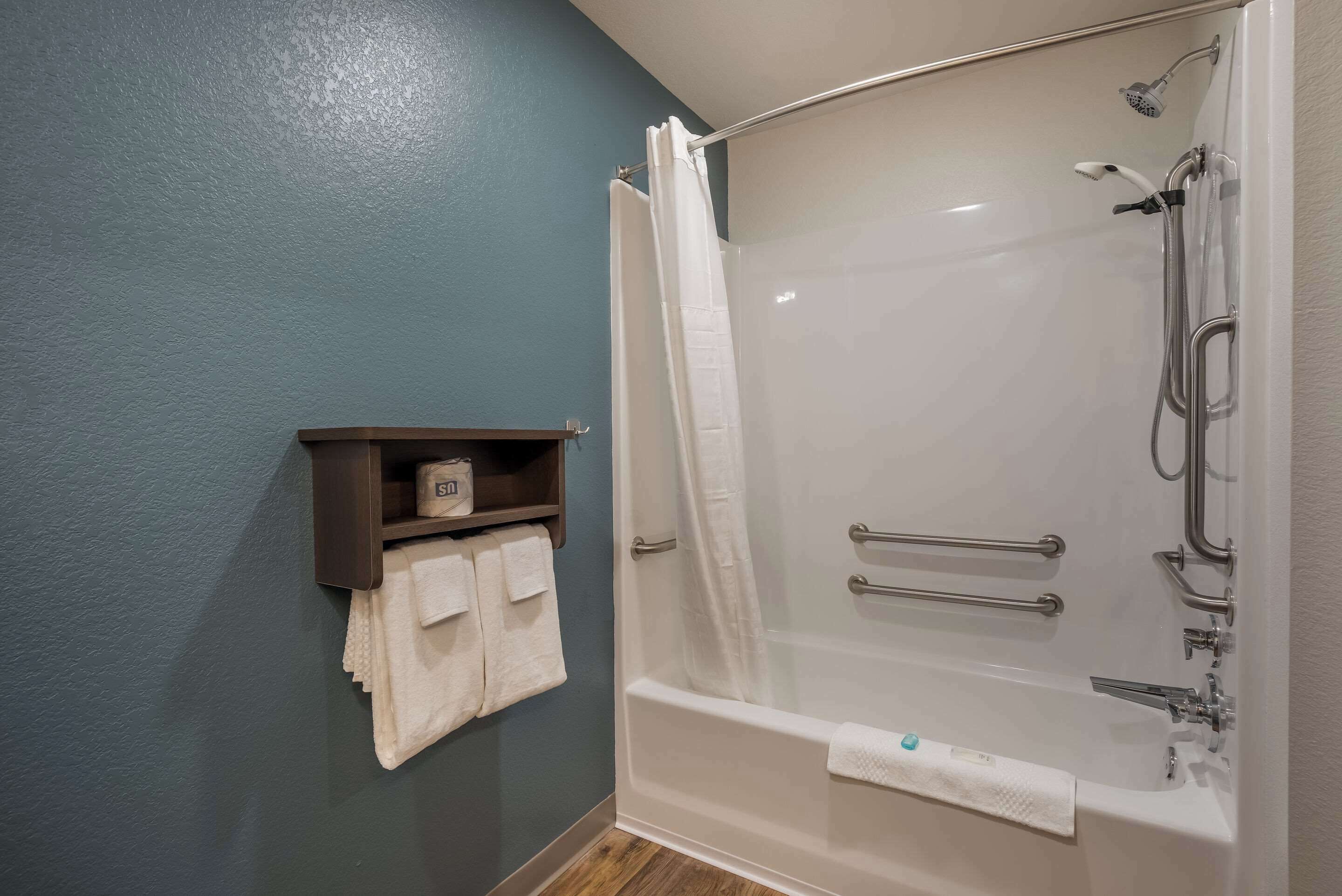 Une salle de bain moderne avec une douche à parois de verre, une étagère en bois où sont posées des serviettes et une baignoire équipée de barres d’appui.