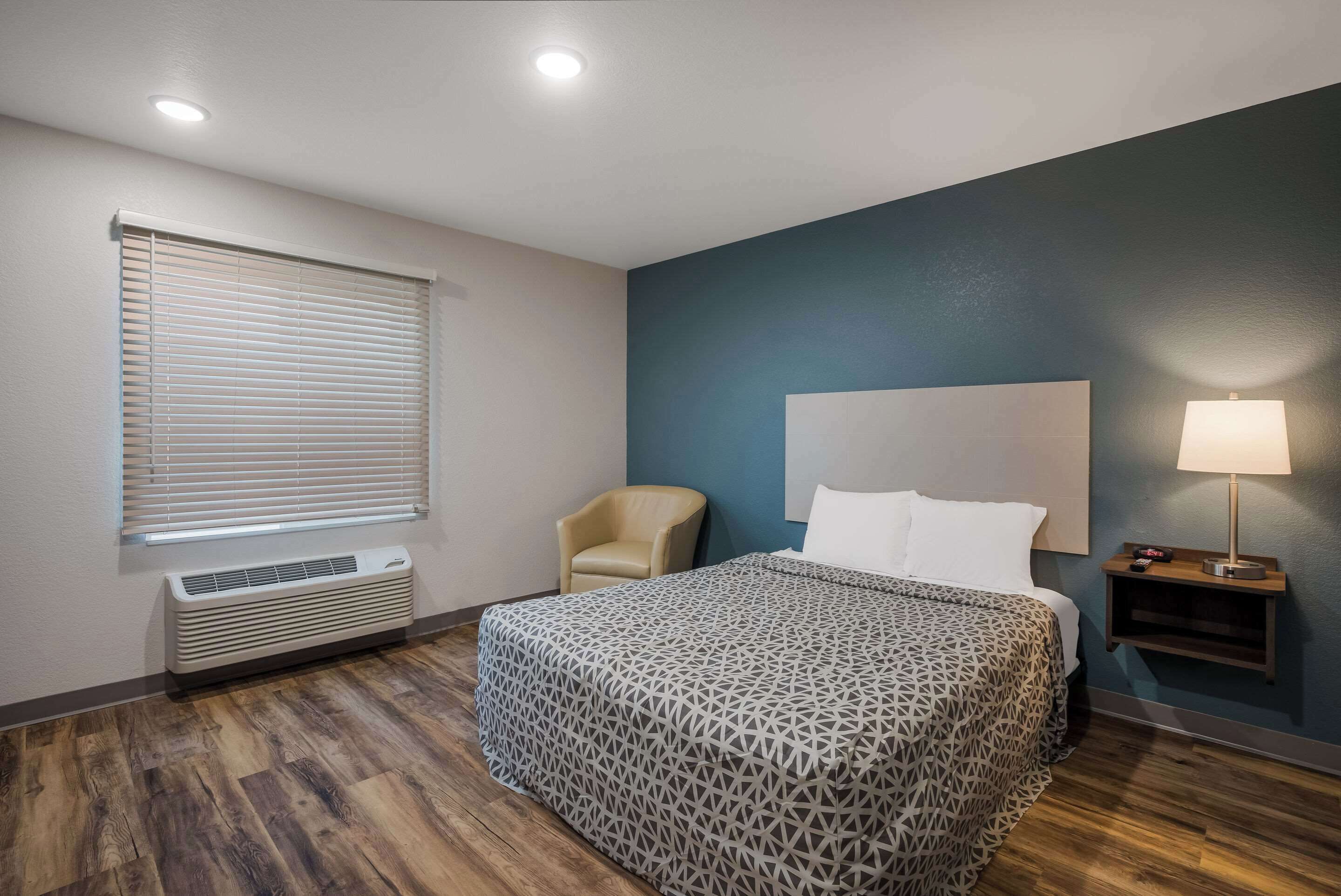 Een hotelkamer met een bed, een stoel, een nachtkastje met een lamp, een raam met jaloezieën en een aan de muur bevestigde airconditioner.