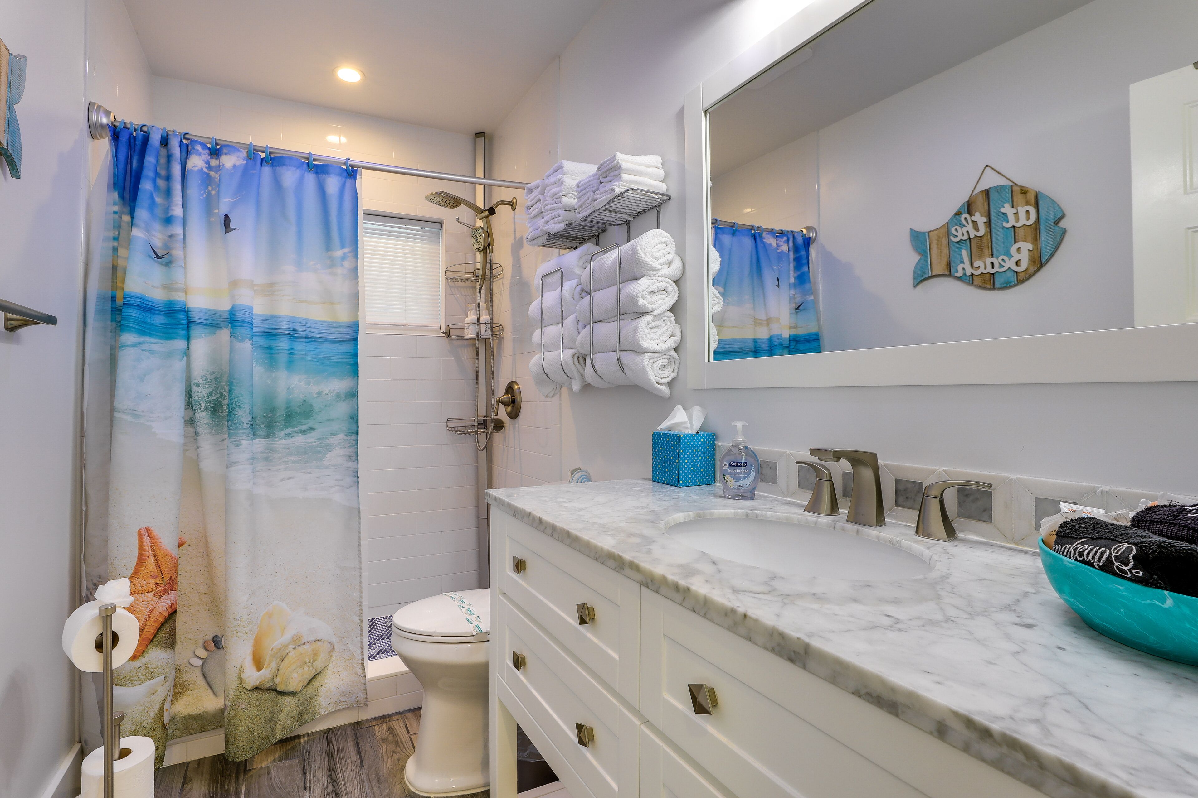 A bathroom with a shower curtain depicting a beach scene, a toilet, a sink with a marble countertop, and a mirror.