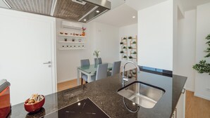 Cozinha moderna com bancada preta, pia de aço inoxidável e forno embutido. Há uma área de refeições com mesa de vidro e cadeiras, além de uma parede branca com prateleiras exibindo objetos decorativos.