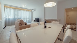 Quarto de hotel moderno com área de refeições, sofá, televisão e uma garrafa de vinho sobre a mesa.