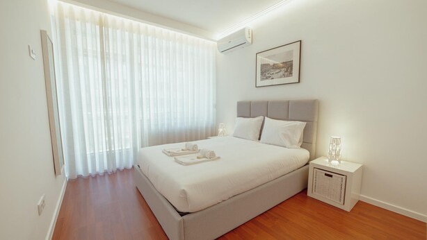 Um quarto com uma cama grande, uma mesa de cabeceira com luminária e uma janela com cortinas leves.