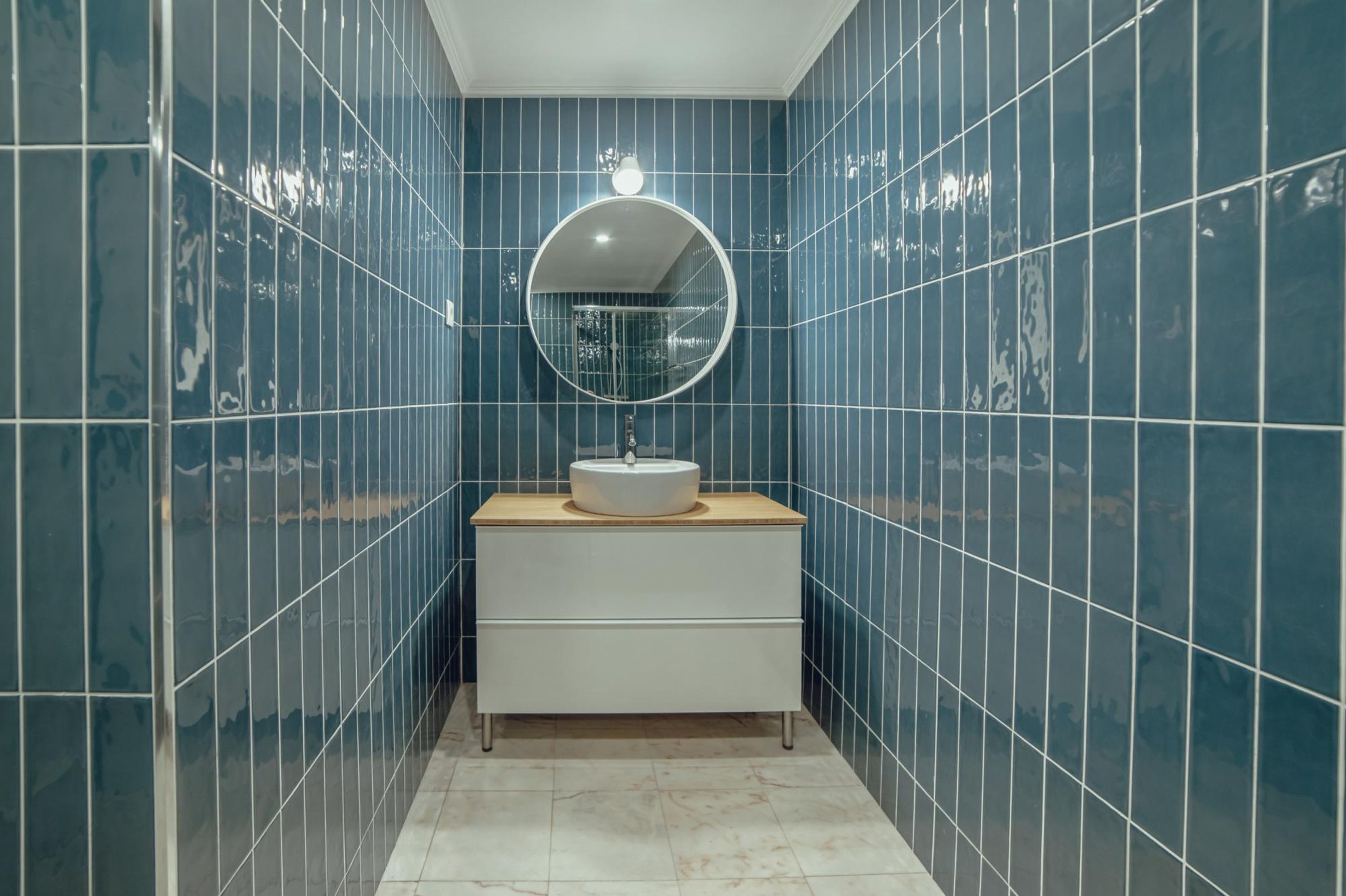 A bathroom with blue tiled walls, a round mirror, a white sink, and a wooden countertop.