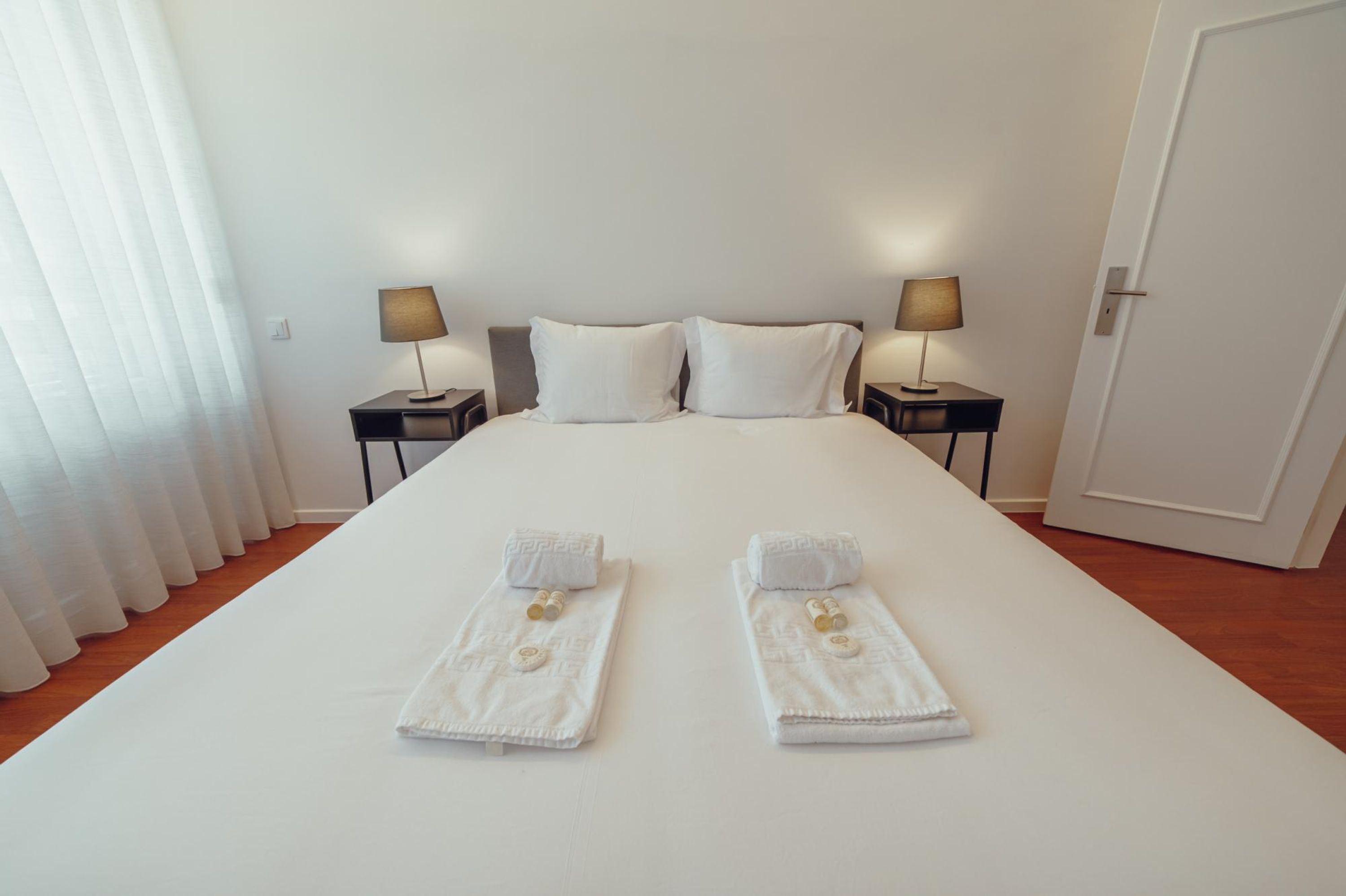 Un lit bien fait, avec du linge de lit blanc, deux tables de chevet avec des lampes et une porte sur la droite.