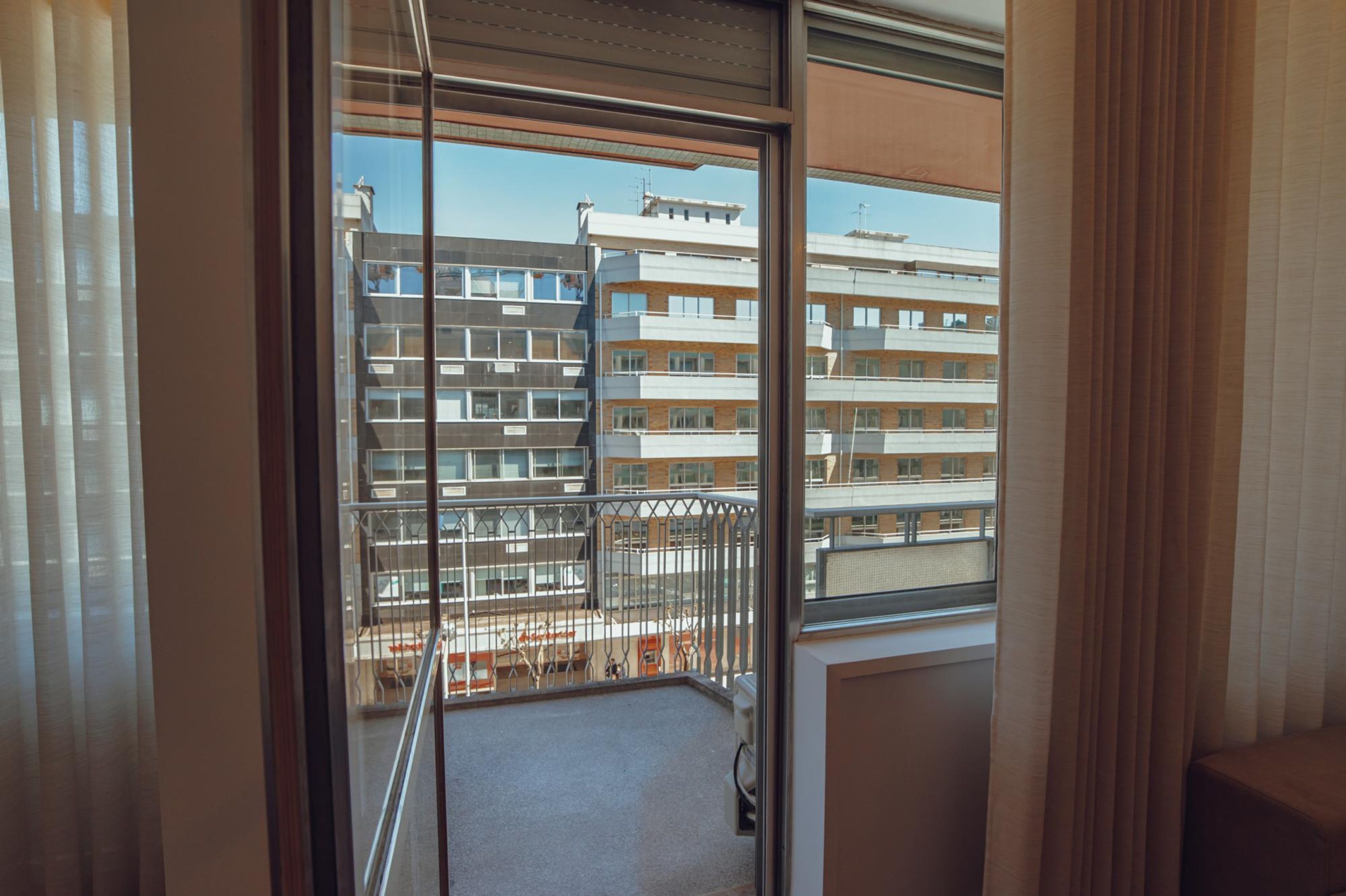 Un balcón con baranda de metal, vista a edificios de varios pisos y una ventana con cortinas.