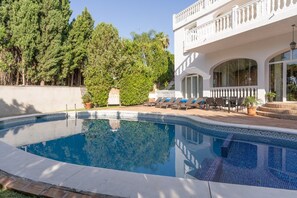 A swimming pool with a white building in the background, a covered patio area, and a view of trees.
