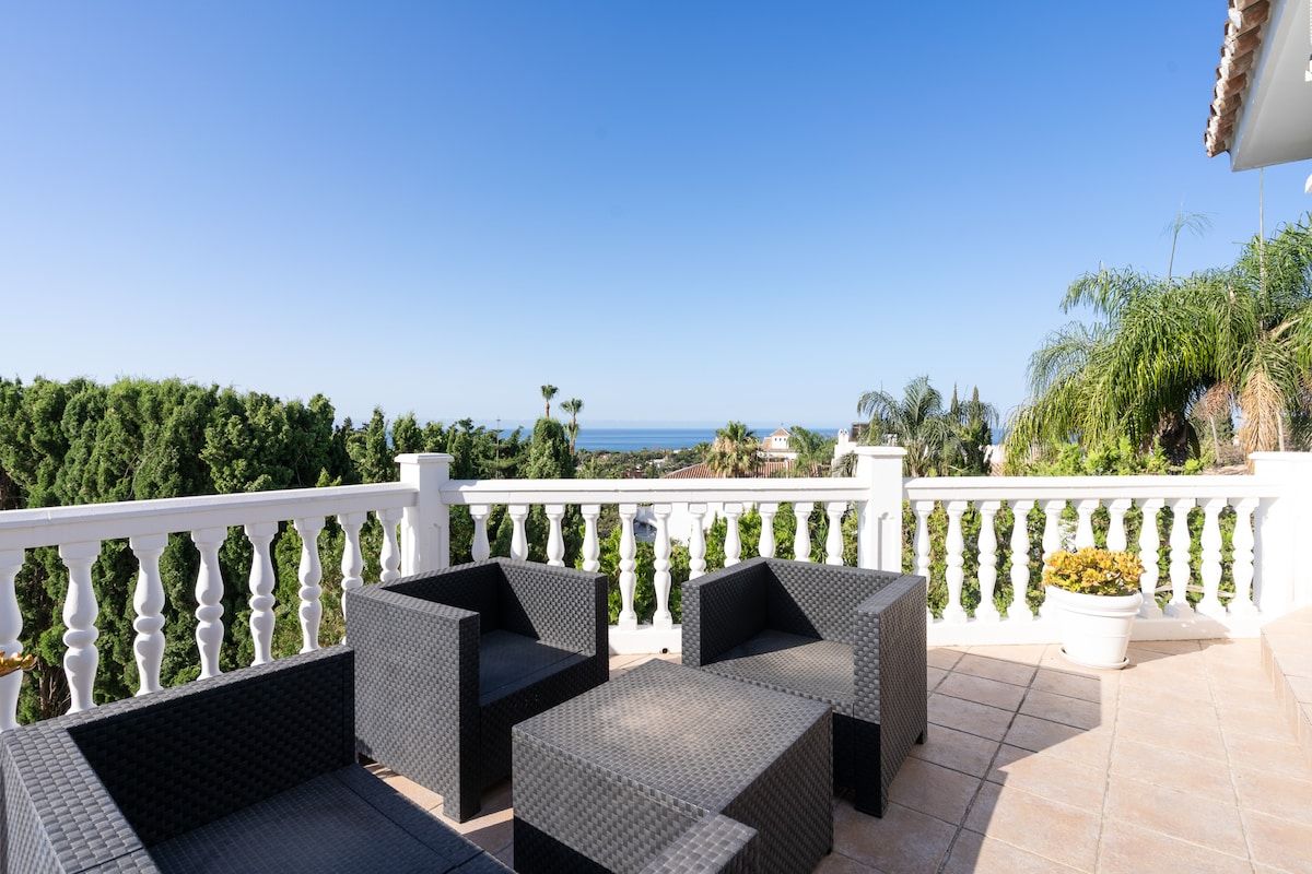 A balcony with wicker furniture and a white balustrade overlooking a coastal landscape.