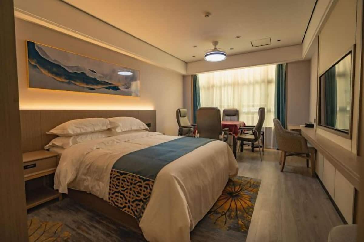 A hotel room with a large bed, a desk, and a chair. There is a painting on the wall and a window with curtains.