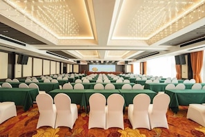 A conference room with rows of tables and chairs, a stage with a podium, and a large screen. - Cotton Rose Hotel (Cili)