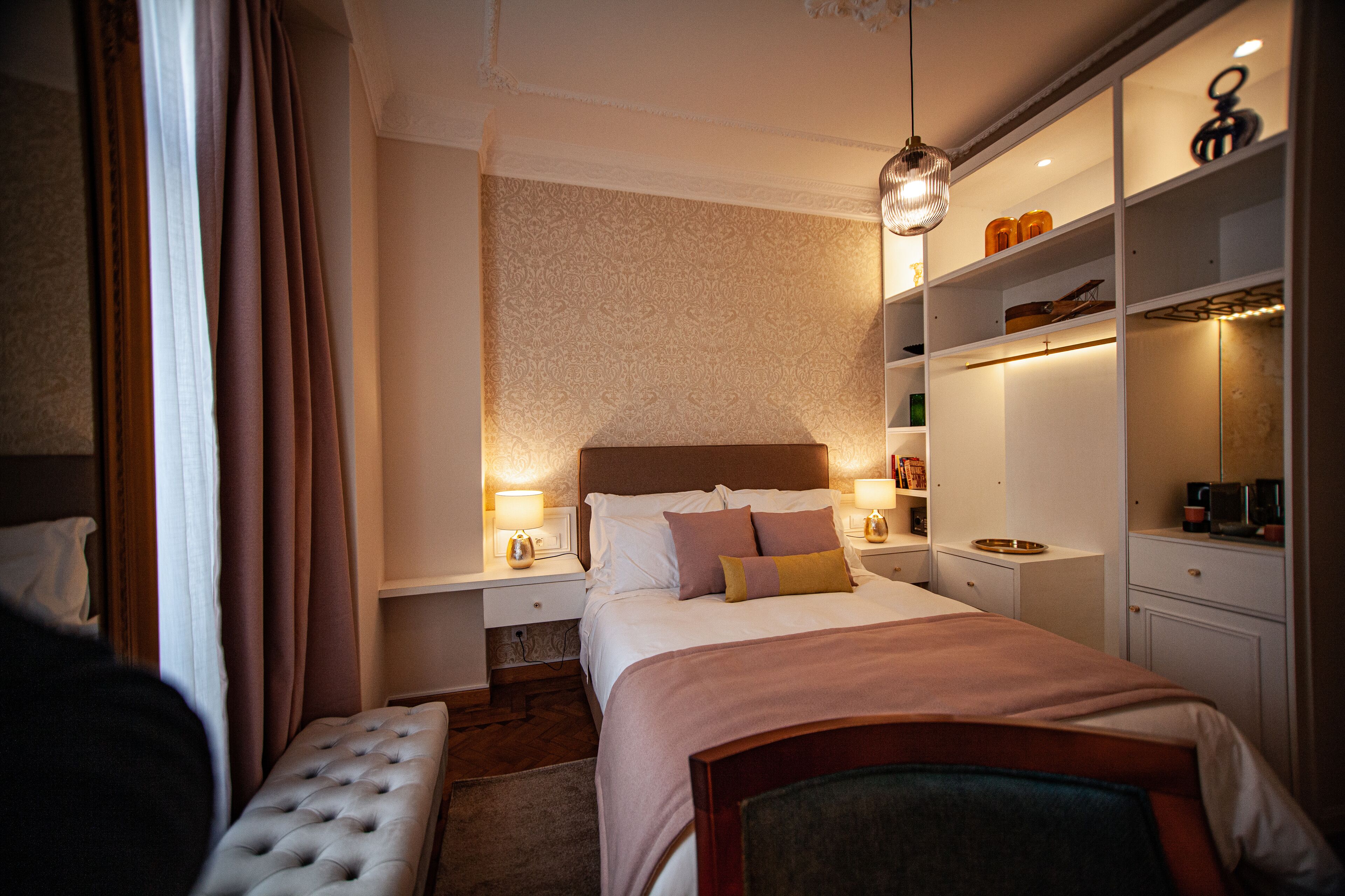 A neatly arranged bedroom with a large bed, bedside tables with lamps, a bench, and a wardrobe with shelves.