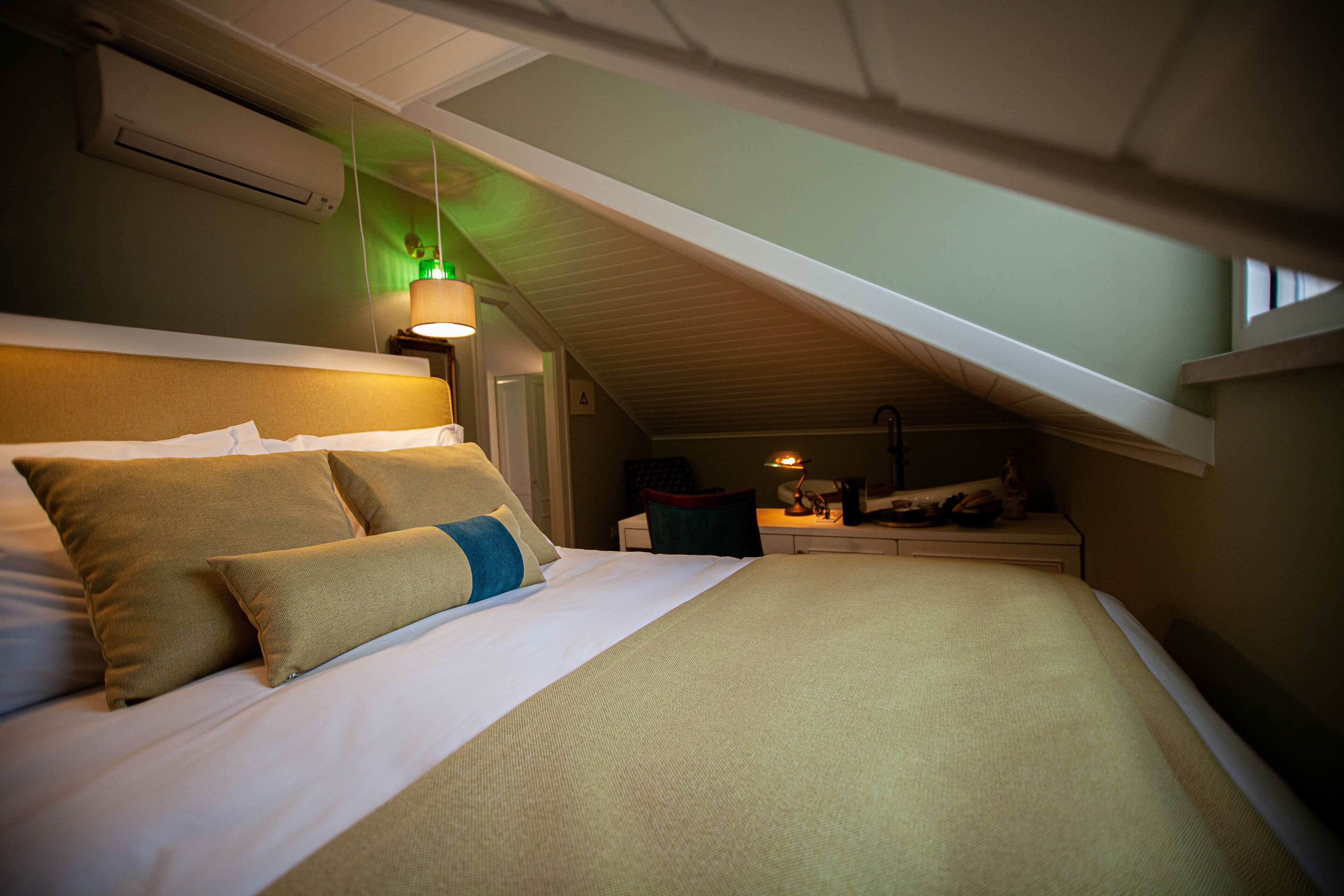 A bedroom with a sloped ceiling, a bed with beige bedding, a nightstand with a lamp, and a window.