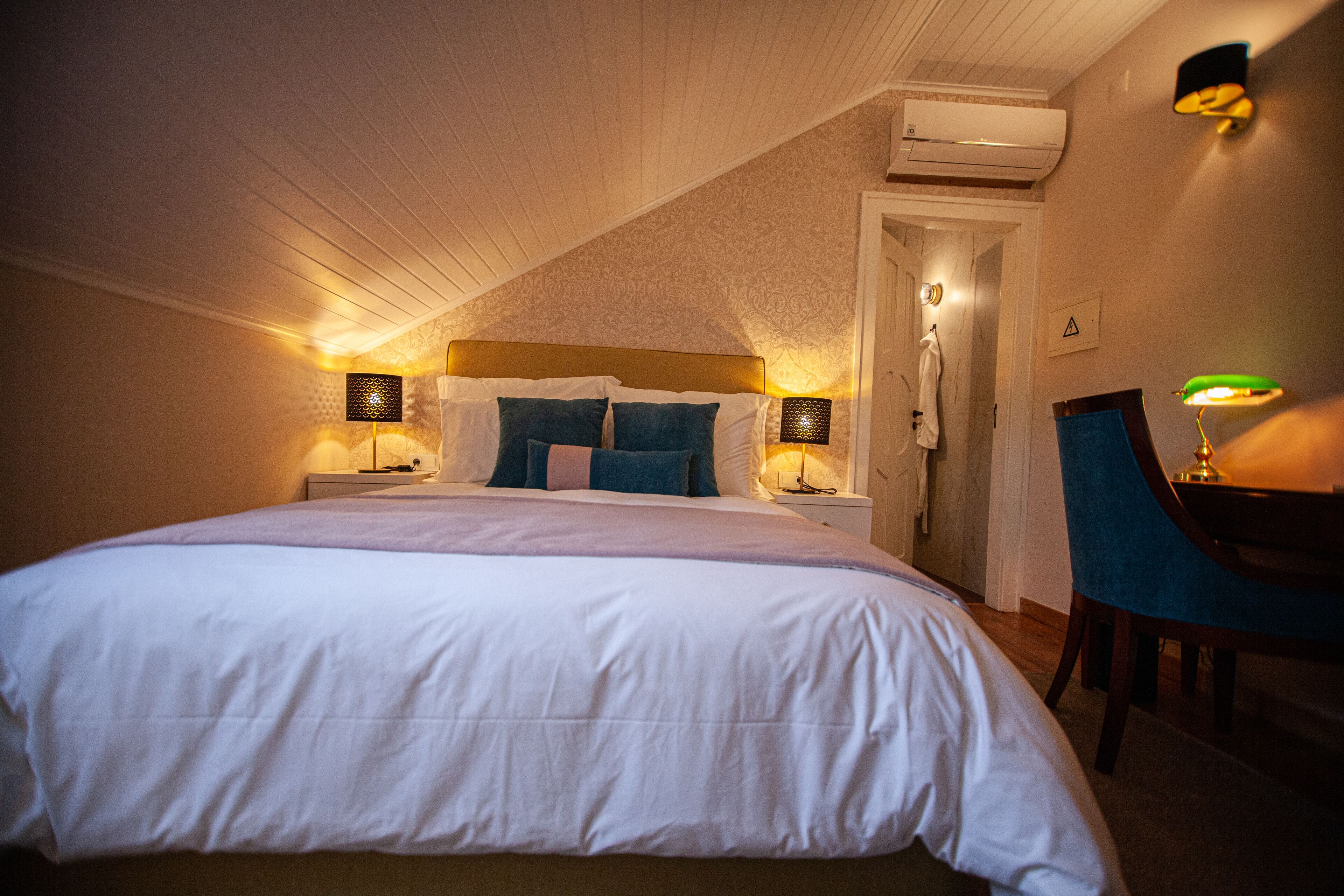 A neatly made bed with white bedding and blue pillows, two bedside lamps, a chair, and an air conditioning unit on the ceiling.