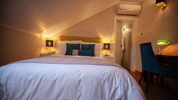 A neatly made bed with white bedding and blue pillows, two bedside lamps, a chair, and an air conditioning unit on the ceiling.