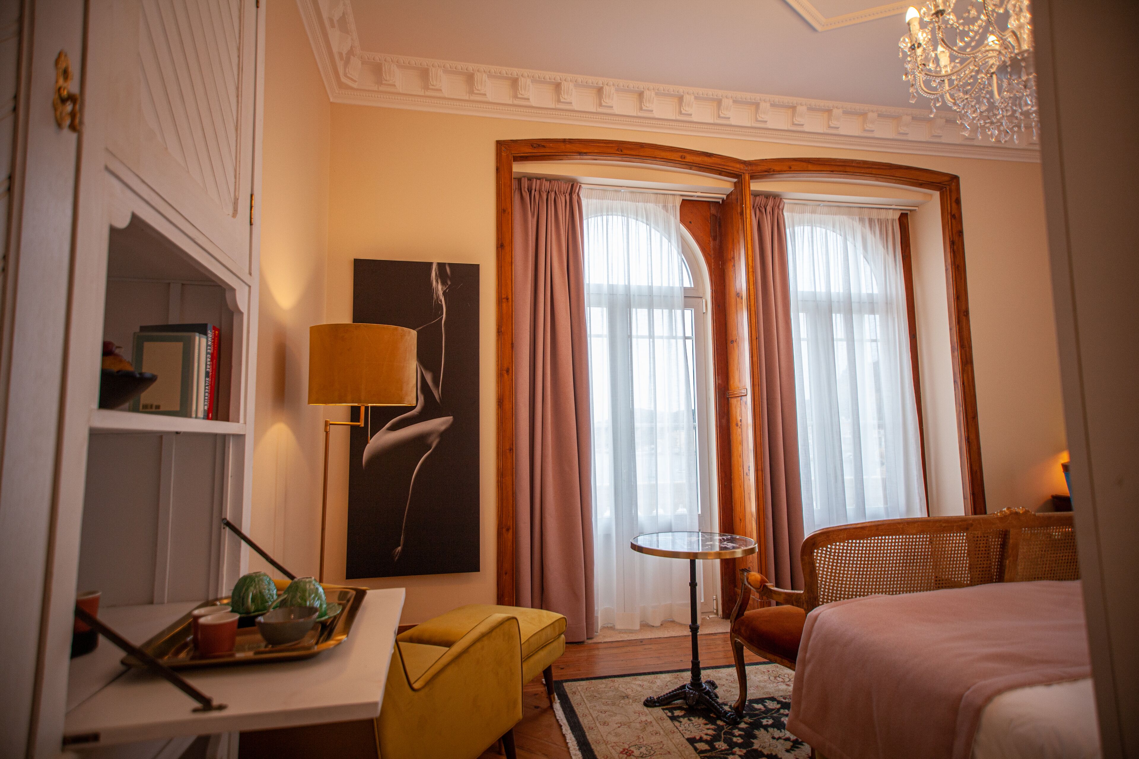 A hotel room with a bed, a desk with a lamp, a chair, a small table, and a bookshelf.
