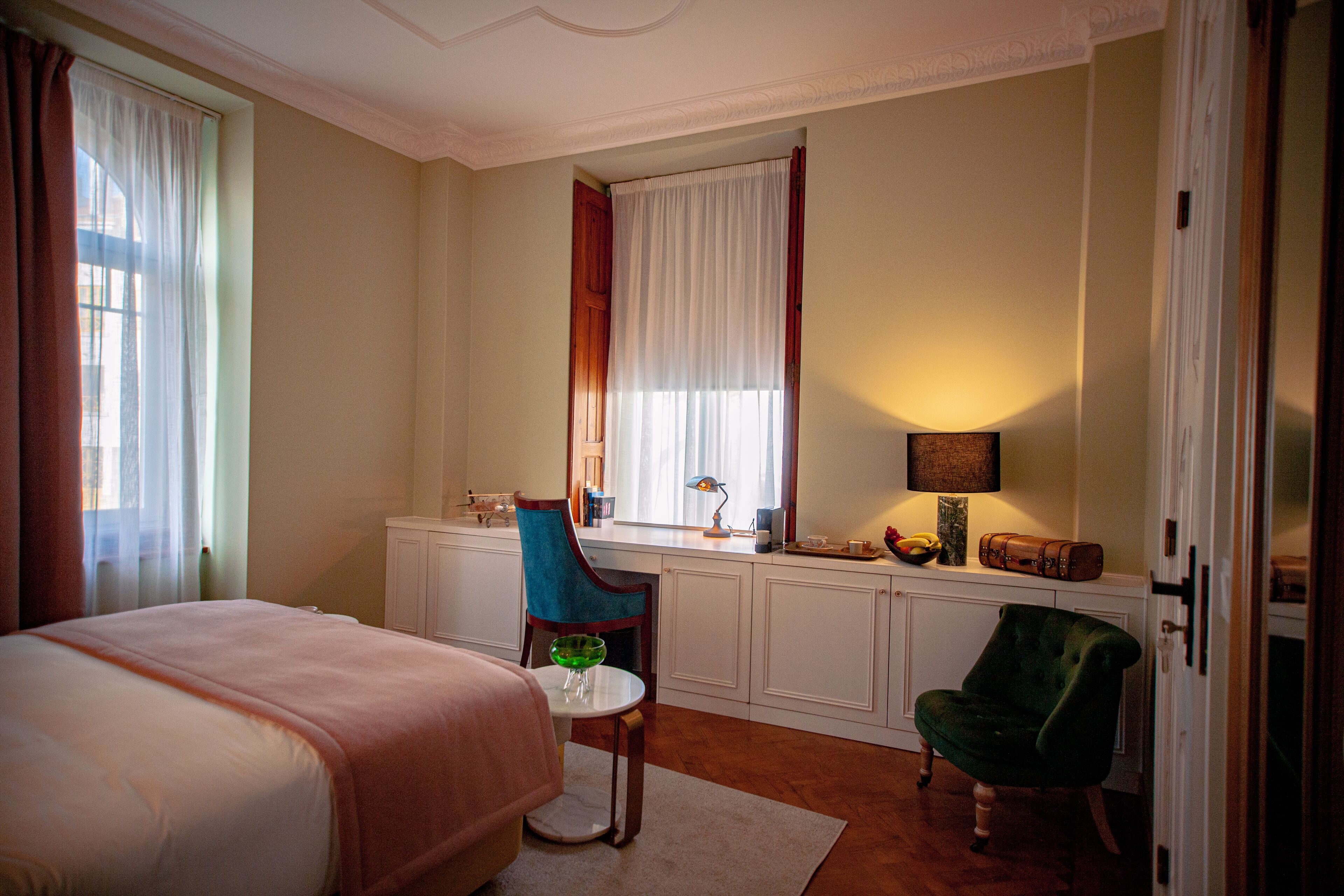 A hotel room with a bed, a desk, a chair, a window with curtains, and a lamp.