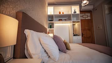Una cama bien hecha con almohadas, una mesita de noche y una lámpara. Se observa un estante con varios objetos y una puerta de madera al fondo.