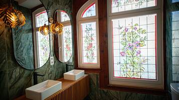 Un baño con pared de mármol, tocador de madera, lavabo blanco, dos espejos redondos y ventana con vitral floral.