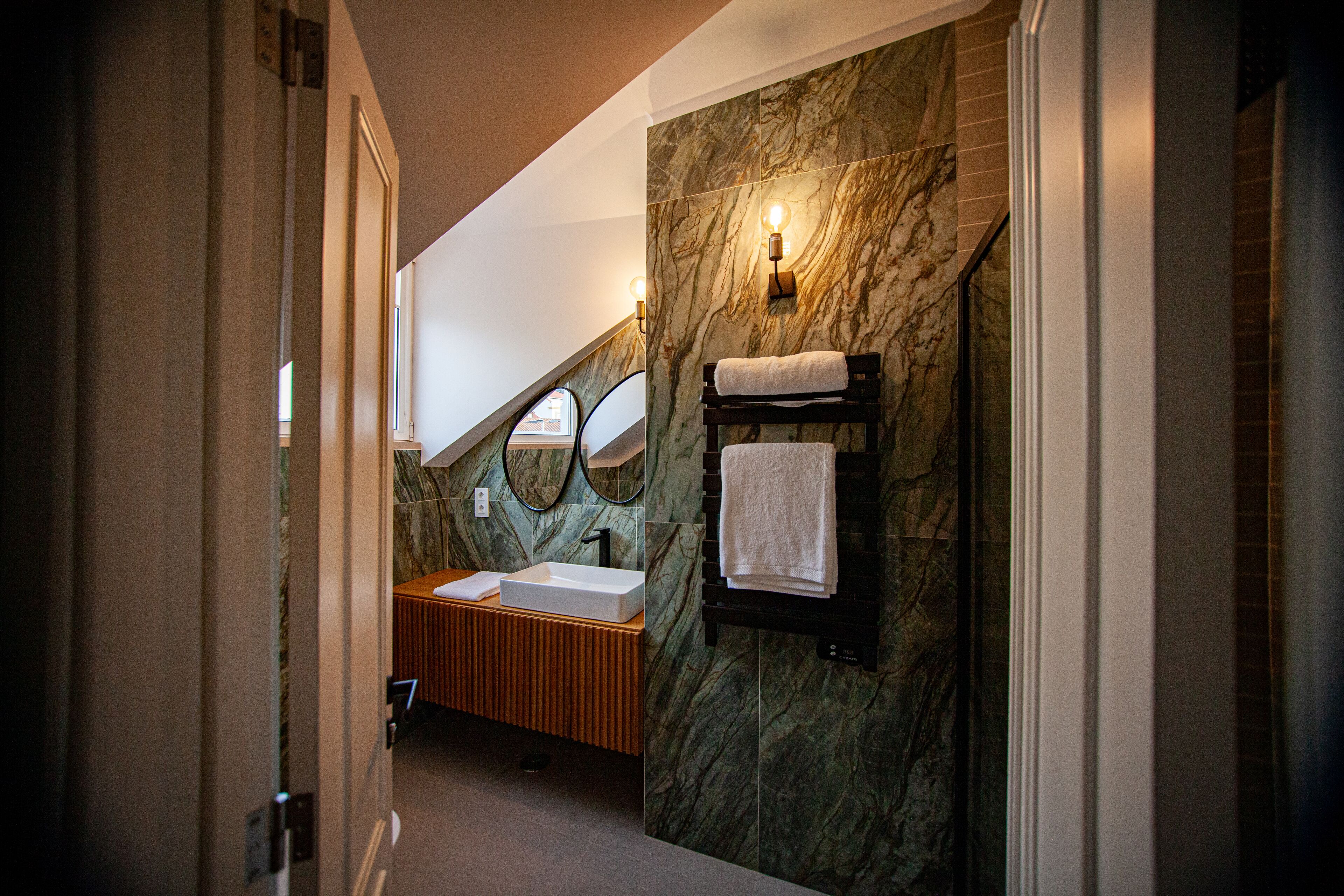 A modern bathroom with a marble wall, a wooden vanity, a sink, a mirror, and a towel rack.