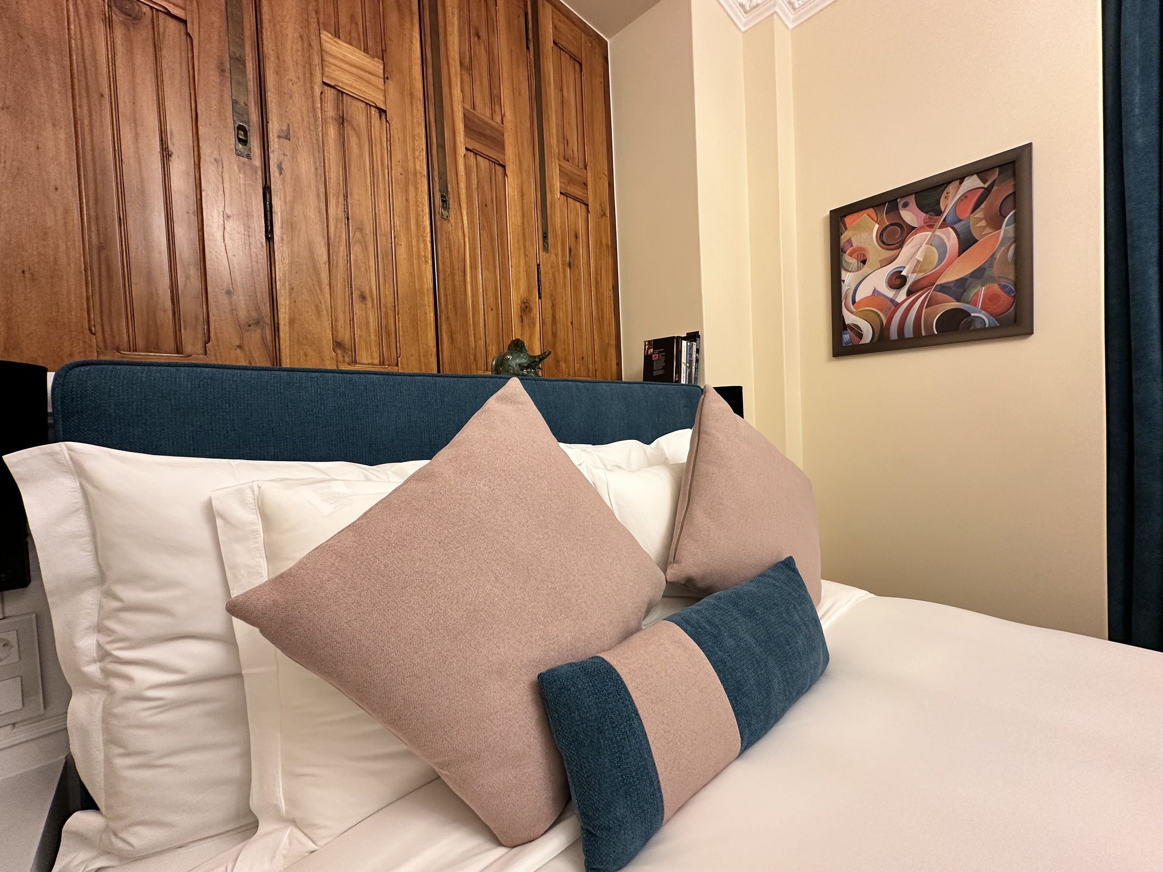 A neatly made bed with a blue headboard, white and beige pillows, and a wooden wardrobe in the background.