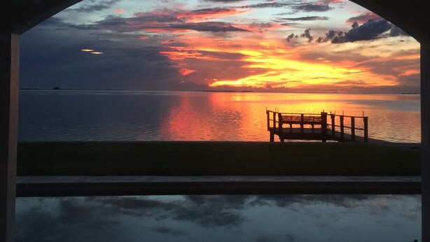 A sunset view through an arched window overlooking a calm body of water with a wooden bench in the foreground. - Enjoy GULF FRONT sunsets in newly renovated private studio in private home. (Palm Harbor)