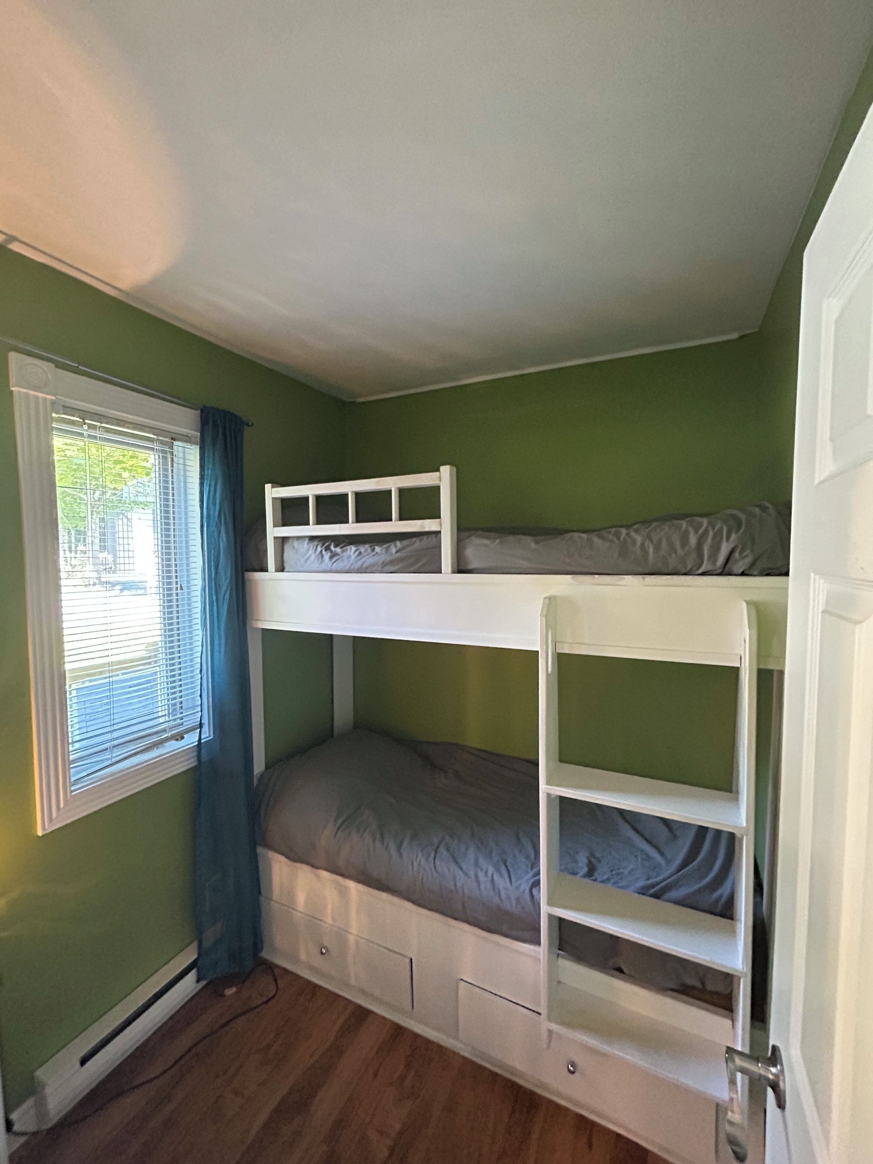Une chambre avec un lit superposé, des murs verts, une fenêtre avec des stores et une porte.
