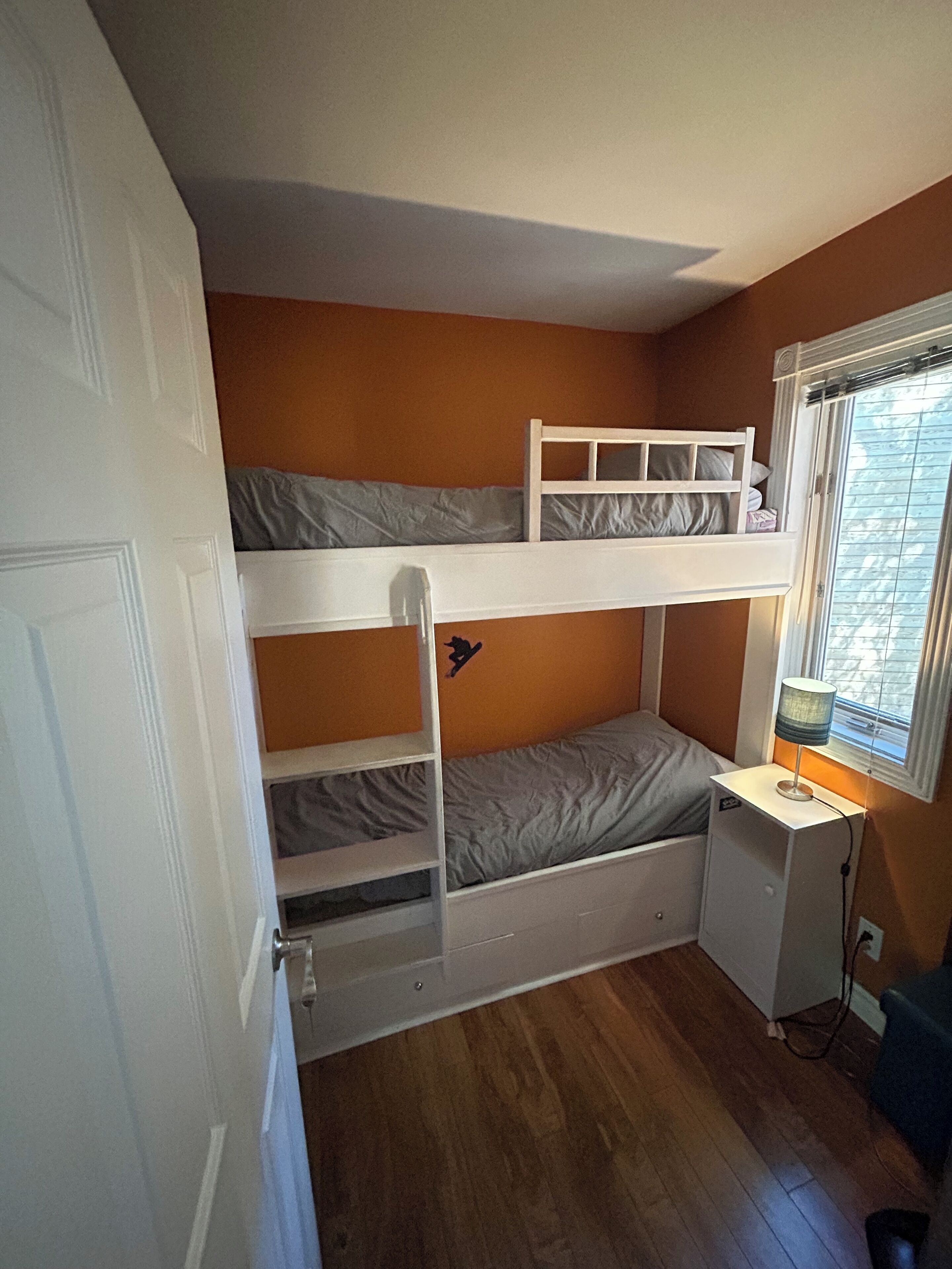 A bunk bed room with a desk and lamp, wooden flooring, and a window with blinds.