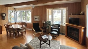 Un salon chaleureux avec une cheminée, des murs en bois, un coin repas avec une table et des chaises, et une grande fenêtre.