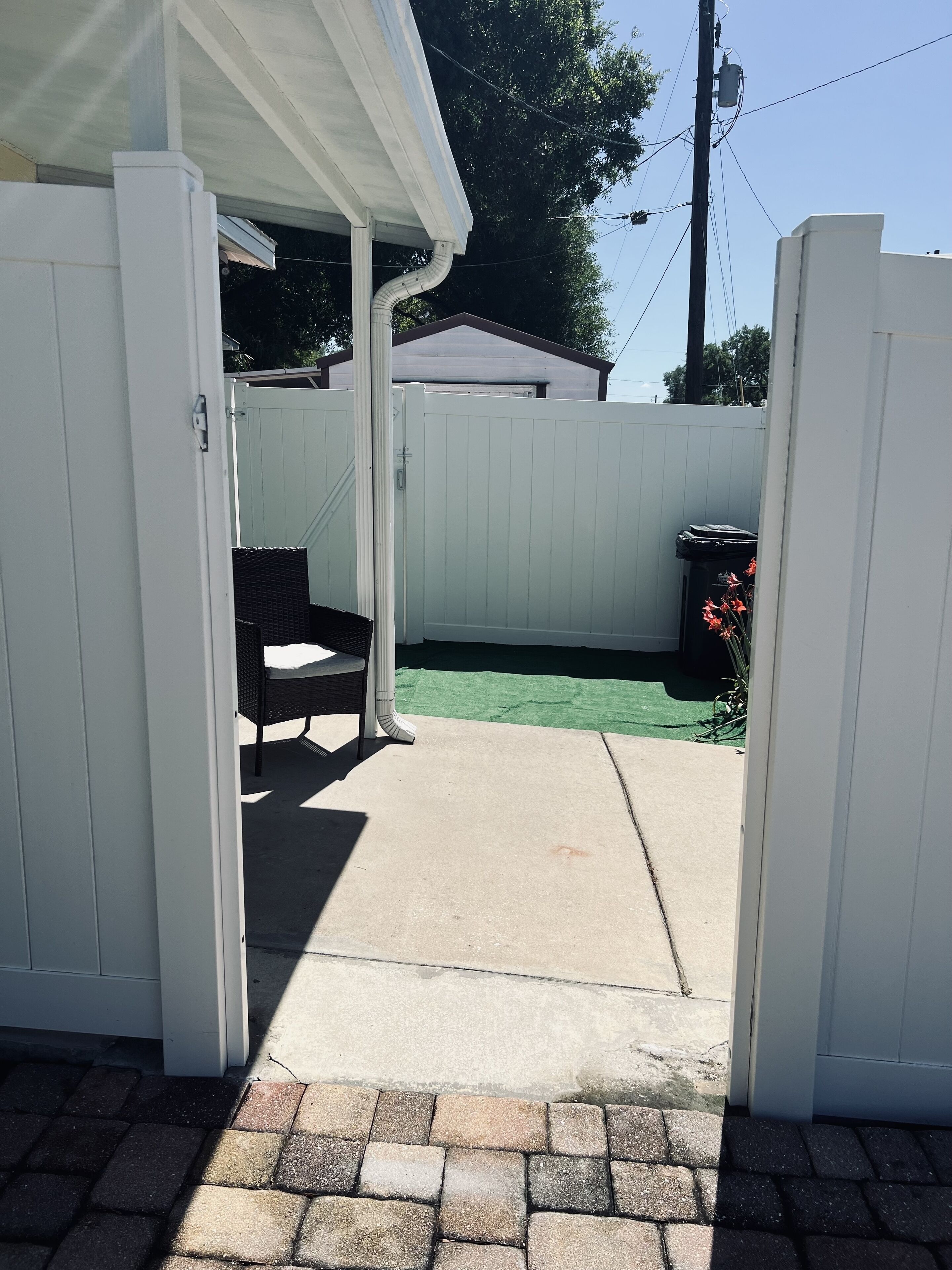 A covered patio area with a concrete floor, a white fence, and a trash bin.