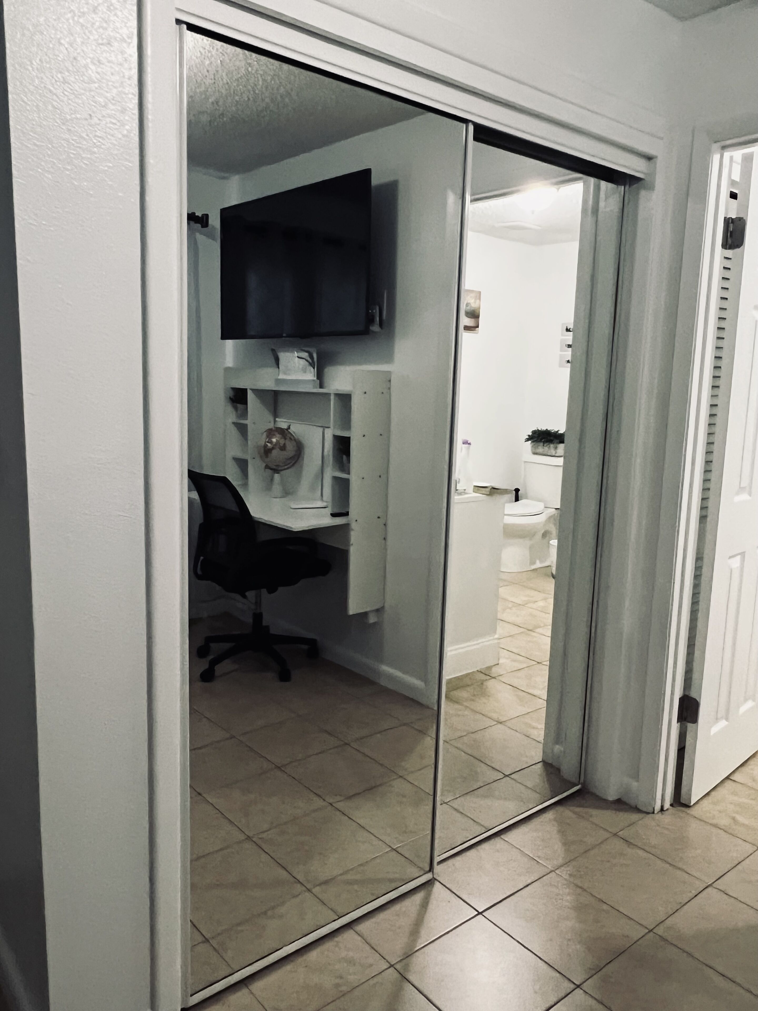 A room with a mirrored closet door, a desk with a chair, a television on the wall, and a toilet visible through the mirror.