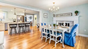 A dining area with a white table, blue chairs, and a chandelier. There is a sign above the fireplace that reads "BE HUMBLE & KIND."