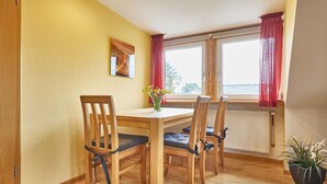 Un coin repas avec des chaises et une table en bois, une fenêtre avec des rideaux, un radiateur et une plante en pot.