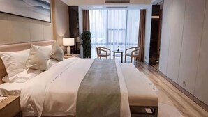 Una habitació d'hotel amb un llit gran, una taula de nit amb una làmpada, una taula petita amb cadires i vistes a l'exterior a través de grans finestres.