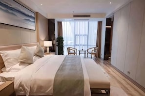 Ein Hotelzimmer mit einem großen Bett, einem Nachttisch mit Lampe, einem kleinen Tisch mit Stühlen und Blick ins Freie durch große Fenster.