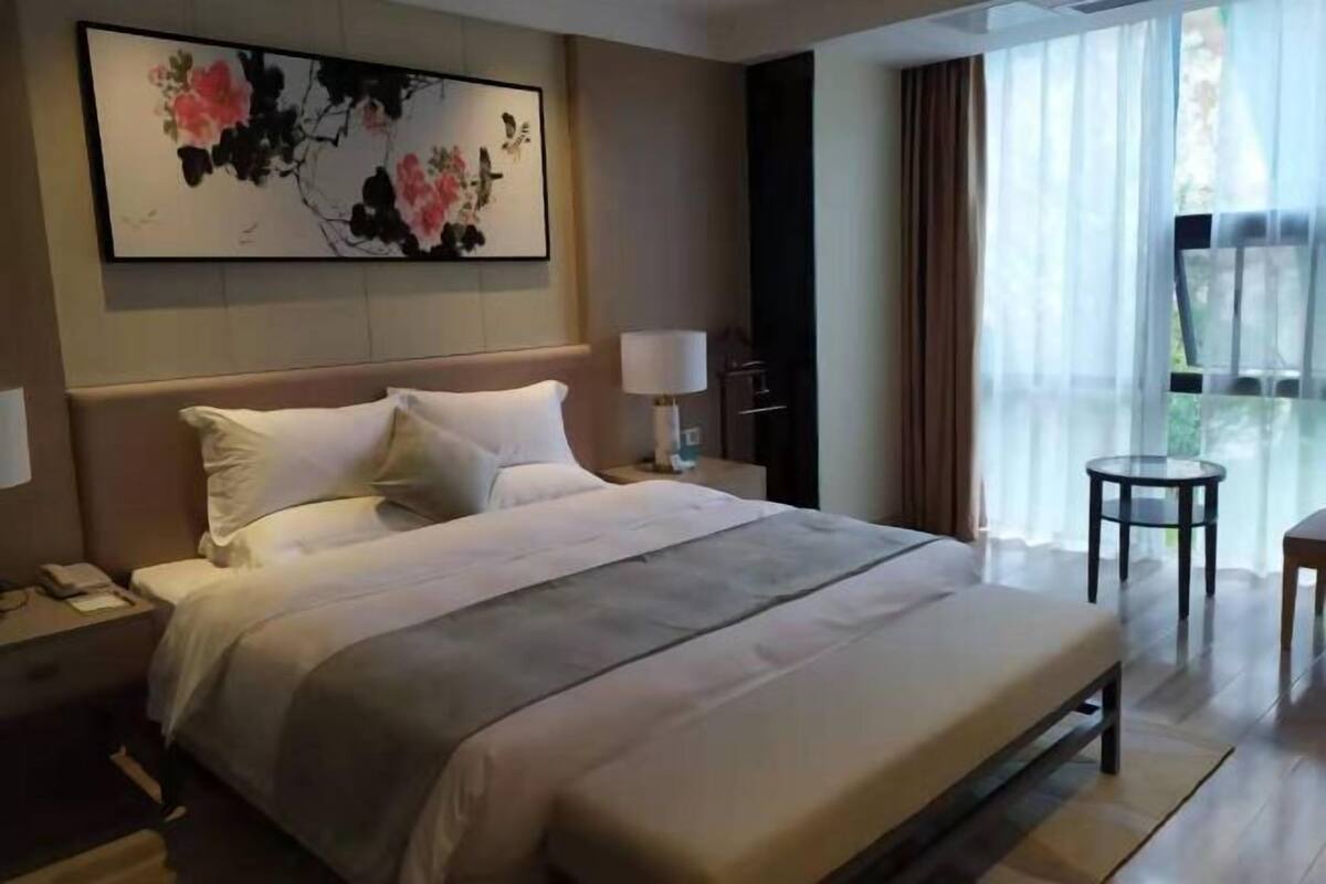 Une chambre d’hôtel avec un grand lit, une table de chevet, une lampe, une petite table et une fenêtre avec des rideaux.