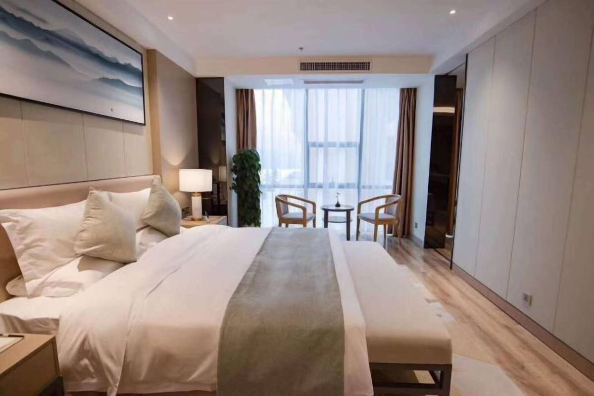 Une chambre d’hôtel avec un grand lit, des tables de chevet, un bureau, deux chaises, une fenêtre avec des rideaux et un tableau au mur.