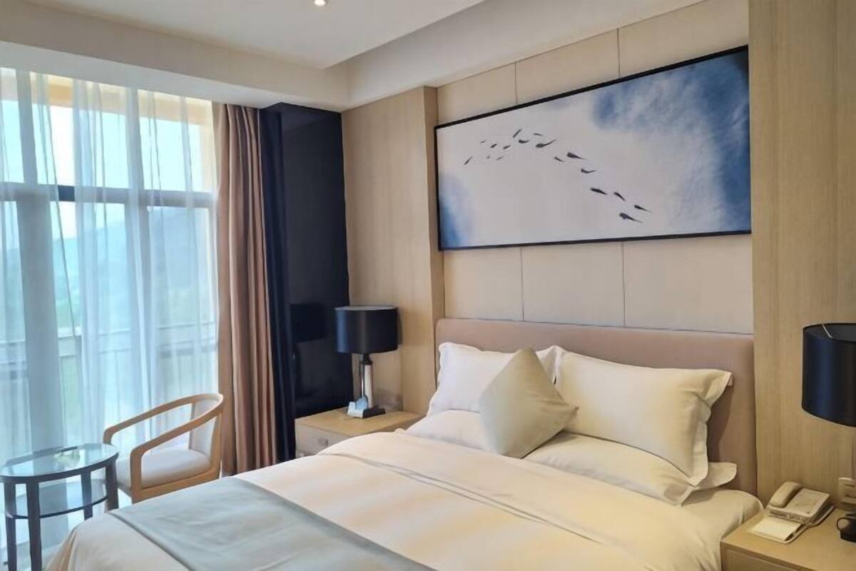 Une chambre d’hôtel comprenant un lit, deux tables de chevet, une chaise, une grande fenêtre avec des rideaux et une œuvre d’art accrochée au mur.