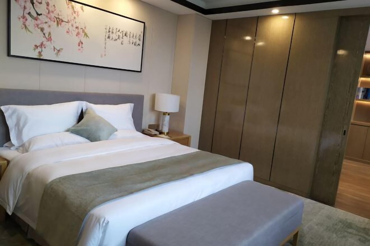 A bedroom with a large bed, a bedside table with a lamp, a wardrobe, and a framed artwork on the wall.